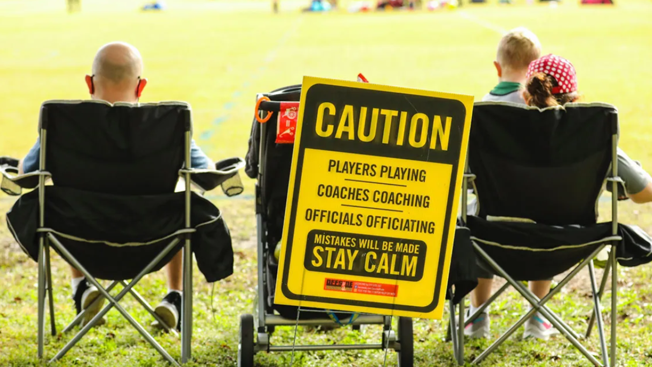 Stock image of youth sports parents in front of a caution sign warning of poor spectator behavior